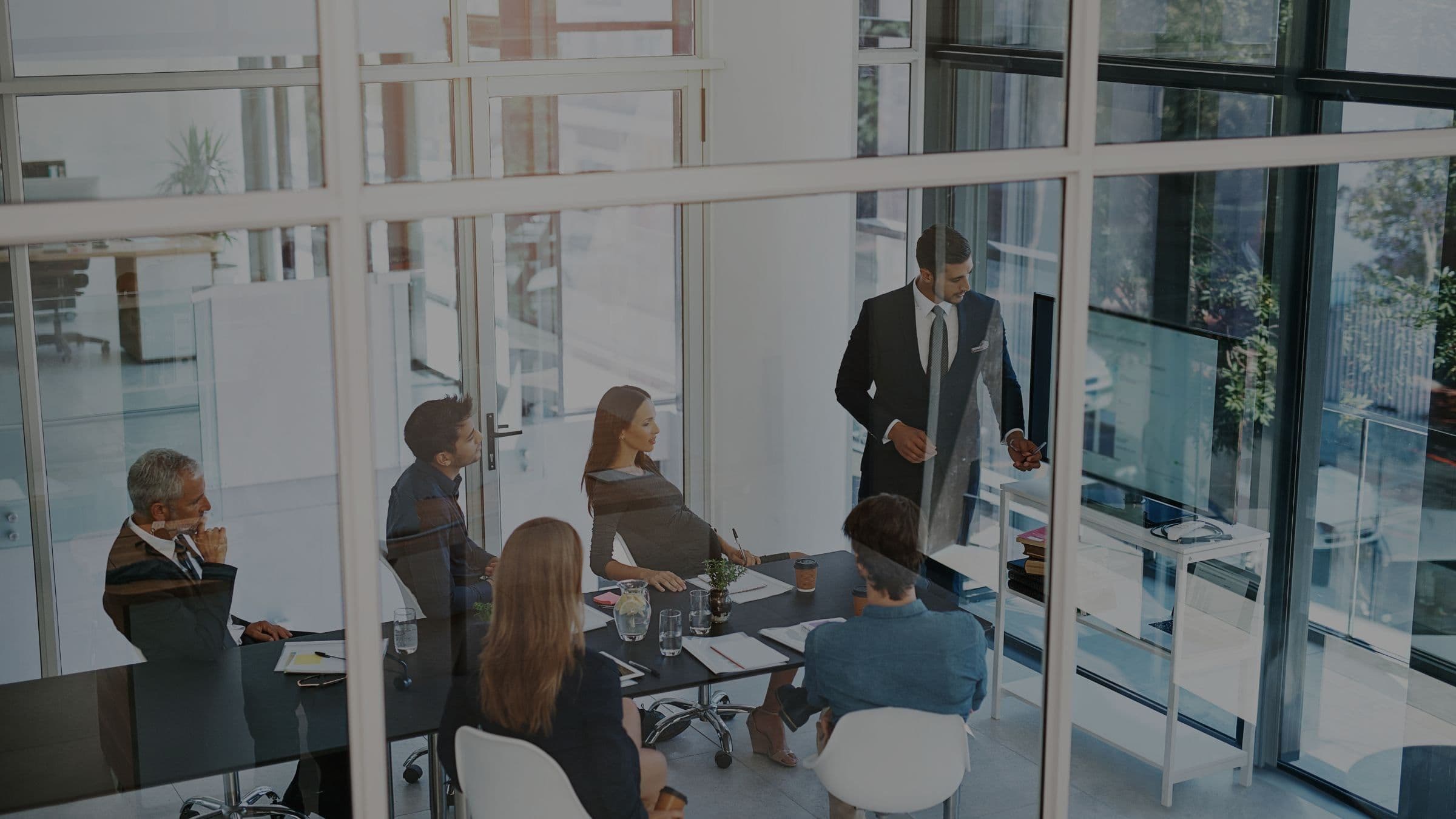 An elevated view through a glazed office wall: five colleagues sit around a dark meeting table while a sixth stands presenting a chart on a wall-mounted screen.