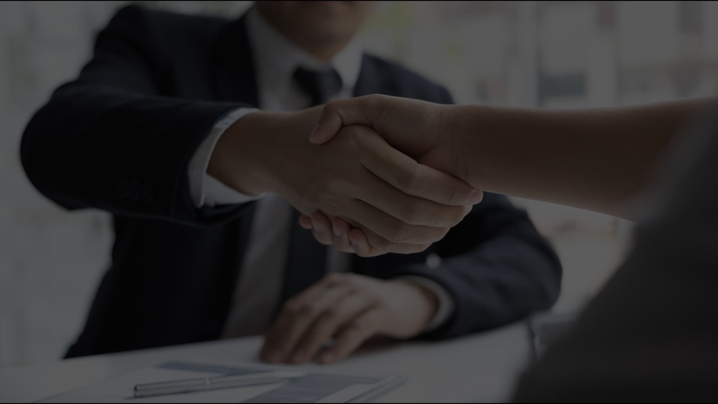 Two people shaking hands across a desk at the close of an interview, with documents and a pen on the table.