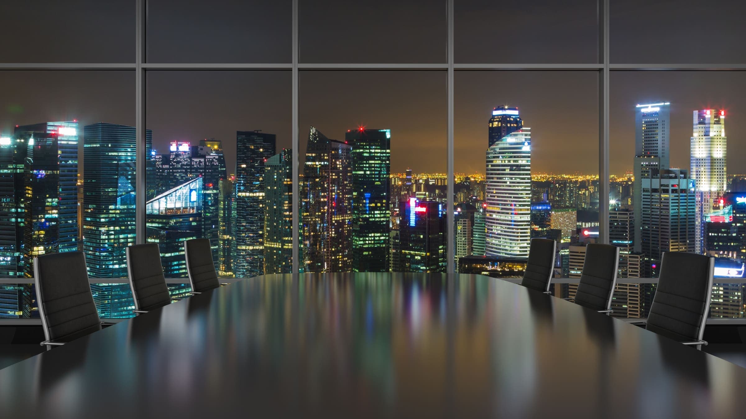 A boardroom table and empty chairs before a window onto a city at night